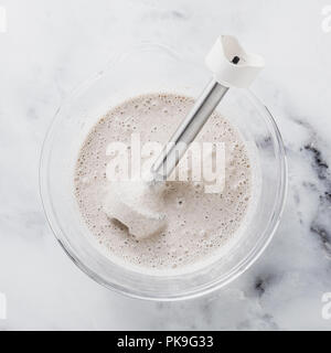 Teig für Pfannkuchen in einer Glasschale mit einem Stabmixer auf einem Marmortisch vorbereiten Stockfoto