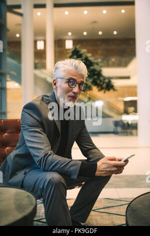 Portrait von Reifen Geschäftsmann in Office Lounge sitzend mit Handy. Ältere männliche Executive in Office Lobby sitzen. Stockfoto