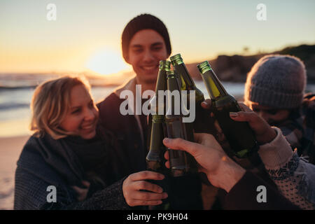 Junge Menschen Toasten mit Bierflaschen am Strand. Gruppe von Männern und Frauen Getränke zusammen am Meer bei Sonnenuntergang. Stockfoto