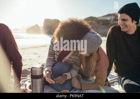 Zwei junge Frau sitzt auf den Strand etwas lustig auf Handy und lächelnd mit einem Freunde sitzen. Gruppe von Jugendlichen auf entspannende
