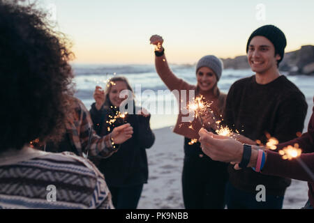 Vielfältige Gruppe von jungen Menschen feiert Tag des neuen Jahres am Strand. Junge Leute Spaß mit Wunderkerzen draussen am Meer. Stockfoto