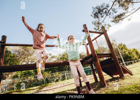 Glücklichen kleinen Zwillingsschwestern Springen eine hölzerne im Park Kleines Mädchen, die Spaß auf dem Spielplatz im Freien. Stockfoto
