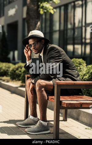 Modische afrikanische amerikanische Mann mit Fedora Hut sitzt auf der Bank Stockfoto