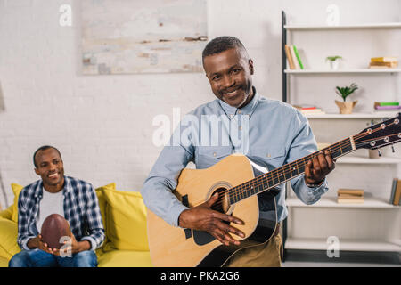 Älterer Mann spielt Gitarre und lächelnd an der Kamera, während erwachsener Sohn holding Rugby Ball hinter Stockfoto