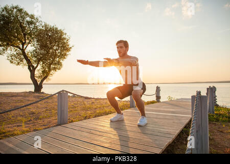 Fitness mensch Ausbildung Luft hocke Übung am Strand vor. Anbringen der männlichen Wahrnehmung crossfit außerhalb. Die Jungen gut aussehenden Kaukasier Männlich fitness Model und Ausbilder im Freien. Stockfoto