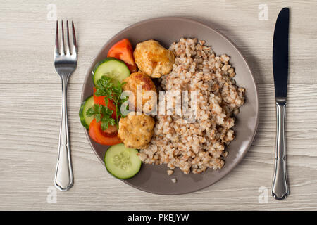 Platte mit Buchweizen, gebratenes Fleisch Schnitzel und Stücke von frischen Gurken und Tomaten mit Zweig der frische Petersilie mit Messer und Gabel auf Holz dekoriert Stockfoto