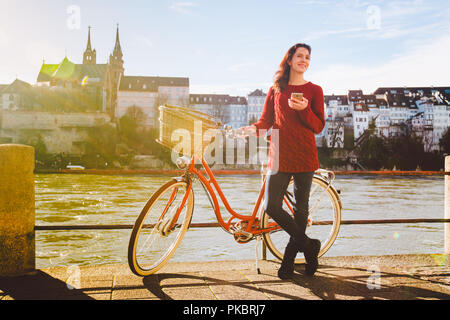 Thema Tourismus auf dem Fahrrad und moderner Technologie. Schönen jungen kaukasischen Frau steht in der Nähe von red retro Fahrrad auf Riverside Rhein Basel Schweizer gewinnen Stockfoto