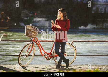 Thema Tourismus auf dem Fahrrad und moderner Technologie. Schönen jungen kaukasischen Frau steht in der Nähe von red retro Fahrrad auf Riverside Rhein Basel Schweizer gewinnen Stockfoto