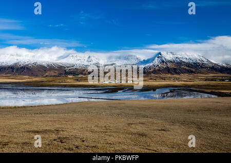 Schnee und bedeckte cloud Berge neben Grasland, Häuser und das Meer bei Leiruvogur Cove, Island Stockfoto