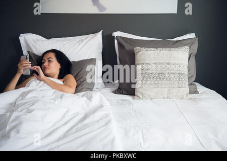 Brunette Mädchen liest Sms von ihrem Freund auf mobilen, während liegt im Bett. Fröhliche junge Frau von Decke bedeckt ist die Wiedergabe von Videos auf Handy vor und nach dem Schlafen. Stockfoto