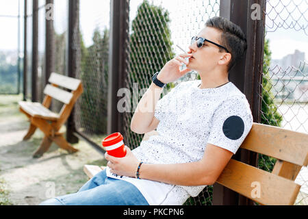 Junge depressiven Mann sitzt auf einer Bank und raucht. Kerl mit einer Tasse Kaffee in der Hand. Stockfoto