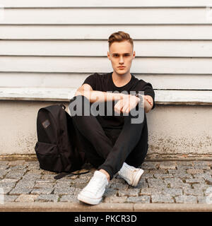 Junge stilvollen Mann im schwarzen T-Shirt und weiße Schuhe mit einem Rucksack sitzt in der Nähe von Holz- wand Stockfoto