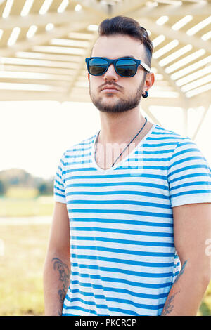 Junge schöne hipster Kerl mit Bart in ein stylisches t-shirt und Sonnenbrille an einem sonnigen Sommertag am Strand. Stockfoto