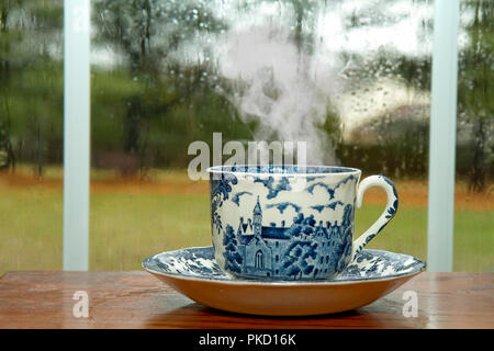 Blau und Weiß-Blaue Tasse Kaffee sitzen am Tisch mit Fenster mit Regentropfen auf Glas als Kulisse. Stockfoto