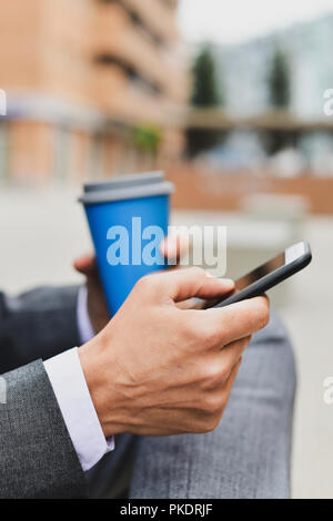 Nahaufnahme eines jungen kaukasischen Geschäftsmann in einem eleganten Grau mit einem Smartphone Anzug und einen Kaffee im Freien Stockfoto