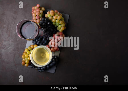 Zwei Weingläser mit Rot- und Weißwein, Flaschen und Trauben auf dunklem Hintergrund. Ansicht von oben. Flach. Platz kopieren Stockfoto