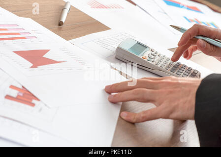 Geschäftsmann Analyse statistische Grafiken mit Hilfe einer manuellen Rechner an seinem Schreibtisch im Büro, in der Nähe Blick auf seine Hände und Schreibarbeit. Stockfoto