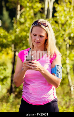 Eine reife Frau tragen aktiv tragen und ein Arm band ihr smart phone Texte, bevor Sie sich für einen Lauf in einem Stadtpark im Herbst Saison zu halten Stockfoto