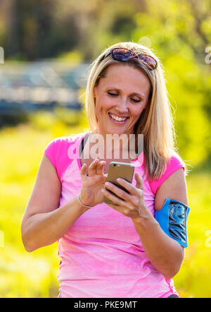 Eine reife Frau tragen aktiv tragen und ein Arm band ihr smart phone Texte, bevor Sie sich für einen Lauf in einem Stadtpark im Herbst Saison zu halten Stockfoto
