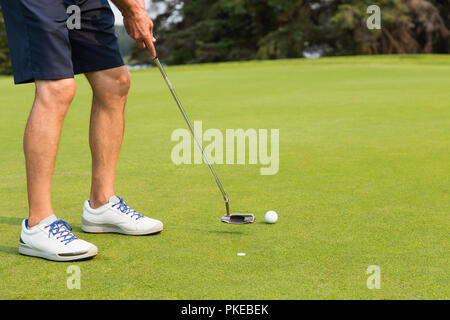 Die Beine, eine reife männliche Golfspieler und Schuhe, als er schwingt auf der Golfball mit einem Putter neben eine Markierung auf dem Putting Grün von einem Golfplatz Stockfoto