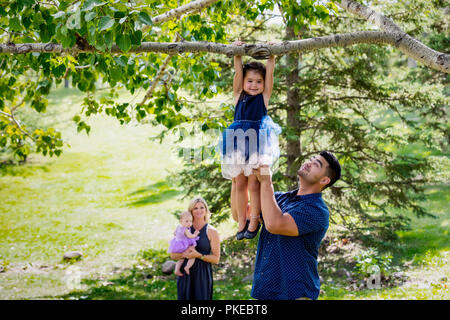 Eine Mutter und eine Babyuhr, während der Vater seine Tochter hoch hält, um ihr beim Aufhängen an einem Ast zu helfen... Stockfoto