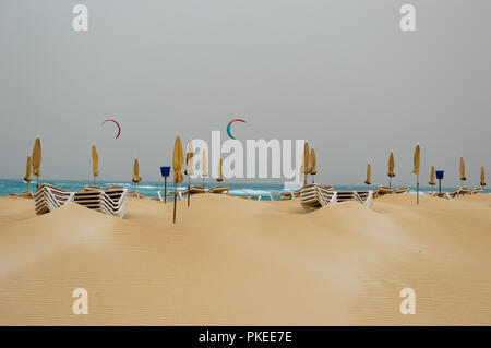 Fuerteventura, Spanien Kanarische Inseln. Gelb gefaltet Sonnenschirm und Sonnenliegen, im Hintergrund sind nur wenige Menschen Kitesurfen Stockfoto