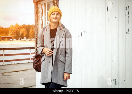 Junge Frau in Blau grauen Mantel, Hut, Jeans zu Fuß entlang der Pier mit einem alten Leuchtturm am Meer entlang. Konzept der Herbstferien an Villag Stockfoto