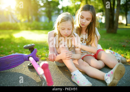 Adorable Mädchen tröstend ihre kleine Schwester, nachdem Sie weg fiel Ihr scooter am Sommer, Park. Kind, der beim Reiten ein Kick scooter verletzt. Aktiven Famil Stockfoto