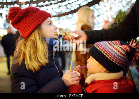 Zwei adorable Schwestern trinken heiße Schokolade und Schokolade essen Pfannkuchen am traditionellen Weihnachtsmarkt. Kind mit Süßigkeiten, Bonbons und gingerbr Stockfoto