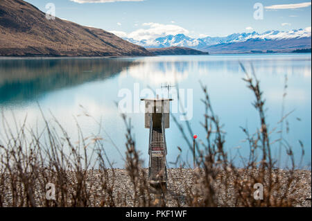 Lake Tekapo, Neuseeland. Küstenlinie mit kleinen elektrischen Unterstation, türkisfarbenen See und Blick auf die schneebedeckten Alpen im Hintergrund. Stockfoto