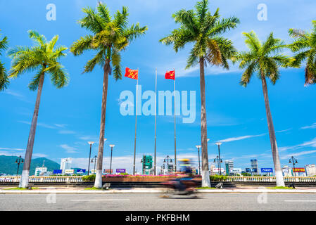 Da Nang, Vietnam - Mai 7, 2018: Mehrere Palmen vor blauem Himmel, Fahnen von Vietnam & Vietnam Kommunistische Partei, Biker, Gebäude der gegenüberliegenden Ufer. Stockfoto