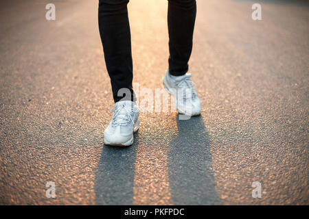 Männliche Füße sind entlang einer Asphaltstraße bei Sonnenuntergang. Weißen Turnschuhen und schwarze Jeans Stockfoto