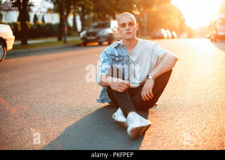 Schöne Jugend junge Mann in stilvollen modische Jeans Kleidung sitzt auf Asphaltstraße bei Sonnenuntergang Stockfoto