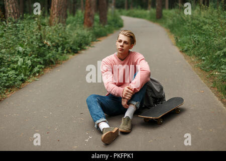 Hübscher junger Mann mit einem Skateboard in einem rosa Pullover und einen Rucksack sitzt auf den Asphalt. Stockfoto