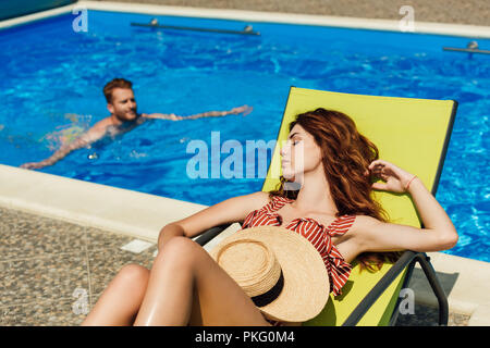 Junge Frau relaxen im Liegestuhl, während ihr Freund schwimmen im Pool auf Hintergrund Stockfoto