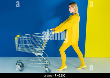 Elegante blonde Frau drücken Warenkorb auf blauem und gelbem Hintergrund Stockfoto