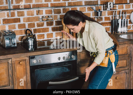 Verwirrt junge repairwoman Suchen in Broken Backofen mit Rauch in Stockfoto