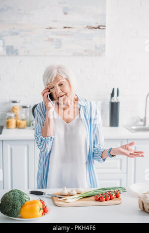 Portrait der älteren Frau im Gespräch auf dem Smartphone beim Stehen an der Theke mit frischem Gemüse auf Schneidebrett in der Küche Stockfoto