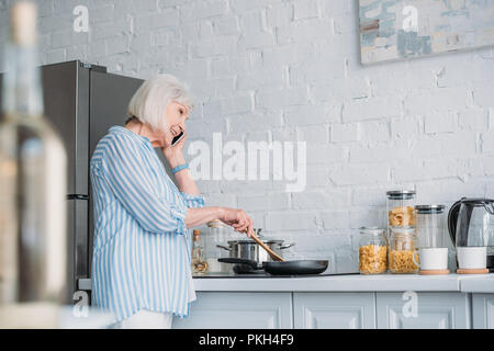 Seitenansicht der lächelnden älteren Frau im Gespräch auf dem Smartphone beim Kochen das Abendessen in der Küche Stockfoto