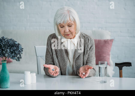 Portrait von grauen Haaren Frau mit Pillen in Händen am Tisch allein zu Hause sitzen Stockfoto