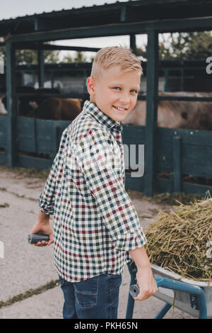 Happy Boy drücken Schubkarre mit Gras und lächelnd an der Kamera im Kuhstall Stockfoto