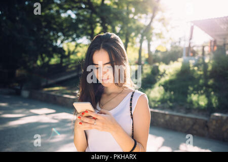 Glückliche junge Frau Brünette mit langen Haaren mittels Smart Phone im Freien während der Spaziergang im Green Park, Sun Flares von hinten. Stockfoto