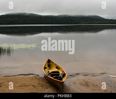 Ein leichtes Kanu mit Fly Fishing Rod. Paddel und Schwimmweste an einem Sandstrand auf einem Adirondack Mountains, NY Wüste See auf einem nebligen Morgen Stockfoto