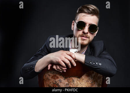 Mann in der Brille sitzt auf einem Stuhl. Unternehmer zuversichtlich Mann. SEO Manager posieren auf einem schwarzen Hintergrund. Erfolgreiche Unternehmer Stockfoto