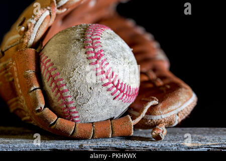 Baseball Handschuh und Ball auf verwittertes Holz Bank. Nahaufnahme mit selektiven Fokus auf die Abgewetzt Textur Detail. Kopieren Sie Platz im schwarzen Hintergrund. Stockfoto