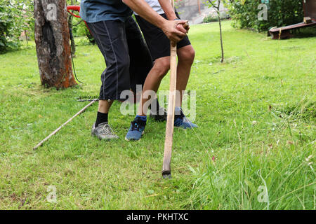 Ein Vater ihm Sohn mit Sense. Ein Mann mit Kind Holding ein Werkzeug und Lehre ein nach rechts verschieben. Sommerzeit Stockfoto