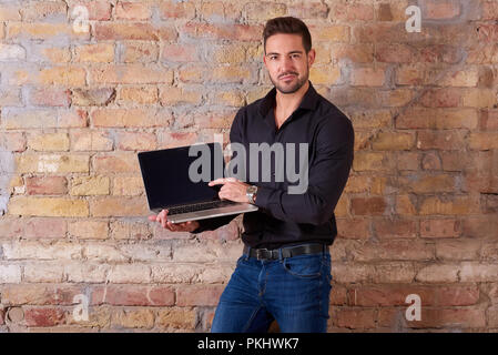 Ein glücklicher hübscher junger Geschäftsmann mit einem Laptop-PC und auf dem Bildschirm in einem schwarzen T-Shirt. Stockfoto