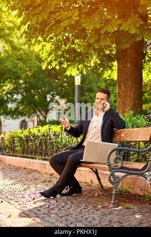Ein hübscher junger Geschäftsmann Standortwahl auf einer Bank mit seinem Laptop an einem sonnigen Straße neben einem Park, der gerade sein Telefon Stockfoto