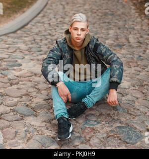 Hübscher junger Mann in stilvollen militärischen Jacke mit Jeans auf dem Boden sitzend Stockfoto