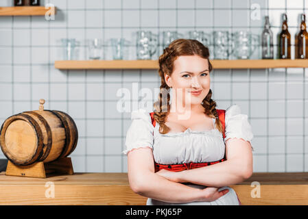 Lächelnd oktoberfest Kellnerin im traditionellen bayerischen Kleid mit verschränkten Armen stand in der Nähe der Theke mit Barrel Bier Stockfoto